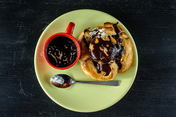 A cup of coffee and a chocolate eclair on a light plate on a black wooden background