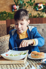 Cheerful Caucasian boy eating a Caesar salad and a burger at a restaurant.