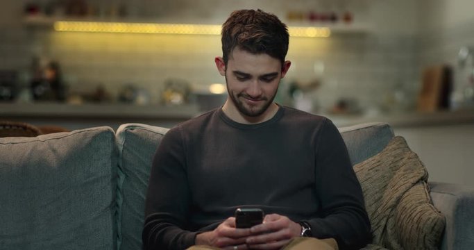 Smiley young man web surfing on his mobile phone on the sofa at home.
