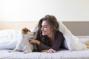 beautiful young woman lying on bed under the white cover with her cute small dog. Home, indoors and lifestyle