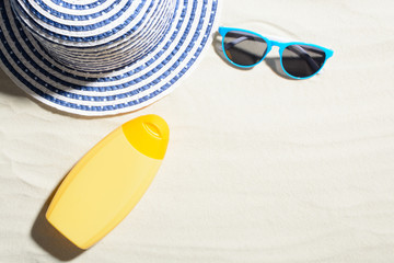 Top view of yellow cream bottle on sand with hat and blue sunglasses