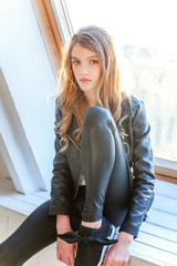 Young teenage girl in leather jacket sitting on modern chair posing in bright light room against white wall background window. Pretty sexy fashion sensual woman dressed in hipster rock style outfit