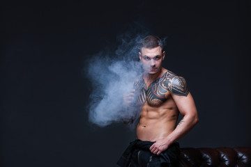 Vaper. The man with a muscular naked torso with tattoos smoke an electronic cigarette on the dark background