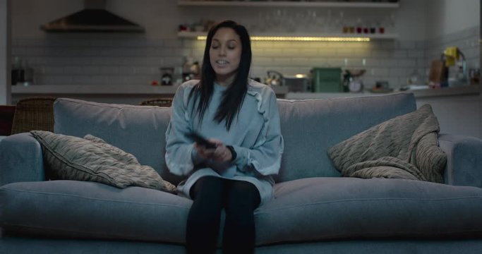 A young attractive woman sits down on a sofa in a darkened living room and switches on the tv.