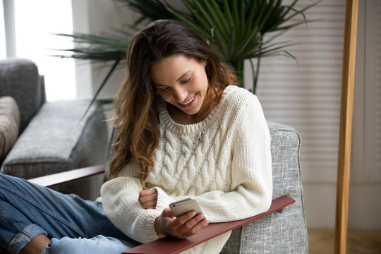 Smiling happy millennial woman holding smartphone relaxing on armchair, teenage girl checking social network news online, reading texting message on cell or using app on new mobile phone at home