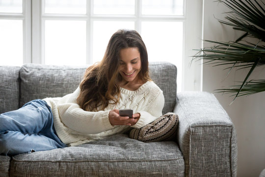Happy young woman lying on sofa at home using smartphone, smiling teen girl holding cellphone enjoying chatting with friend or new mobile apps, texting message or checking social network news online