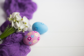 Two colorful Easter eggs and bouquet of tender snowdrops on purple fabric on white wooden table