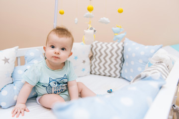 Fototapeta na wymiar Cute baby sitting in a white round bed. Light nursery for young children. Toys for infant cot. Smiling child playing with mobile of felt in sunny bedroom.