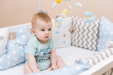Cute baby sitting in a white round bed. Light nursery for young children.  Toys for infant cot. Smiling child playing with mobile of felt in sunny bedroom.
