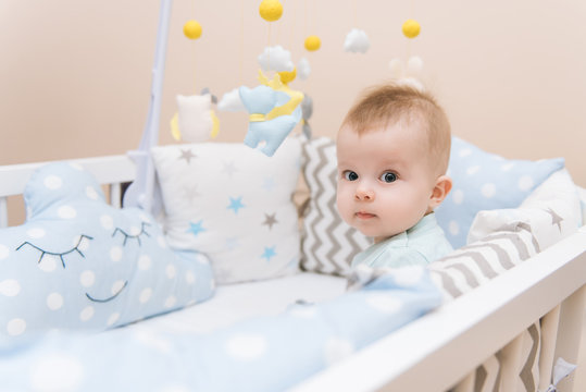 Cute Baby Sitting In A White Round Bed. Light Nursery For Young Children.  Toys For Infant Cot. Smiling Child Playing With Mobile Of Felt In Sunny Bedroom.