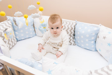 Cute baby sitting in a white round bed. Light nursery for young children.  Toys for infant cot. Smiling child playing with mobile of felt in sunny bedroom.