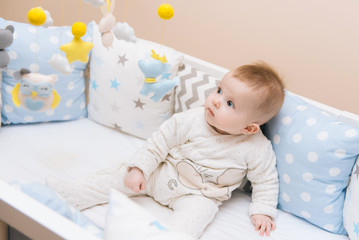 Cute baby sitting in a white round bed. Light nursery for young children.  Toys for infant cot. Smiling child playing with mobile of felt in sunny bedroom.