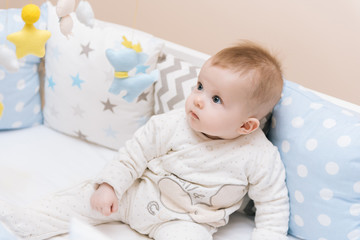 Cute baby sitting in a white round bed. Light nursery for young children.  Toys for infant cot. Smiling child playing with mobile of felt in sunny bedroom.
