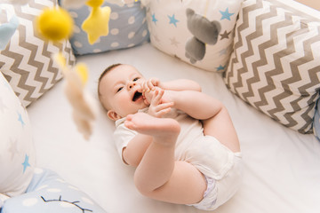 Cute baby lies in a white round bed. Light nursery for young children.  Toys for infant cot. Smiling child playing with mobile of felt and chewing his feet in sunny bedroom.