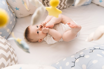 Cute baby lies in a white round bed. Light nursery for young children.  Toys for infant cot. Smiling child playing with mobile of felt and chewing his feet in sunny bedroom.