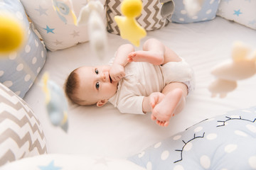 Cute baby lies in a white round bed. Light nursery for young children.  Toys for infant cot....