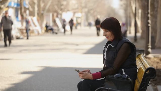 Profile of a smart woman with a bob haircut sitting on a bench and browsing the net to look at photos in the sunny alley in spring