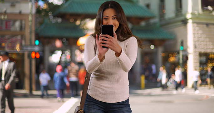 Portrait of cheerful Latina woman taking photo with cellphone in Chinatown