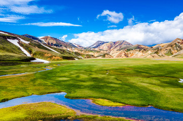 A wide view of Landmannalaugar, one of the most beautiful places in Iceland. A flat green field crossed by streams is surrounded by breathtaking yellow rhyolite mountains