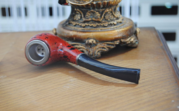 Cachimbo vermelho com tra&ccedil;os pretos, pousado numa mesa junto do p&eacute; de um cadeeiro em madeira trabalhada 