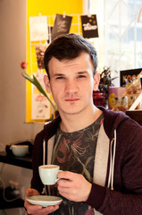 young guy trying to drink espresso on the palate in a coffee house holding a cup and a plate looking at the camera
