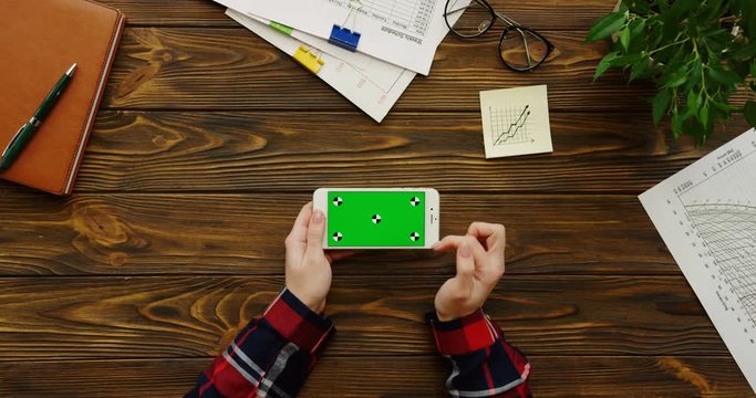 Close up of the female hands with the red plaid sleeves using the white smartphone with a green screen horizontally on the wooden office table with office stuff and plant. Chroma key. Top view
