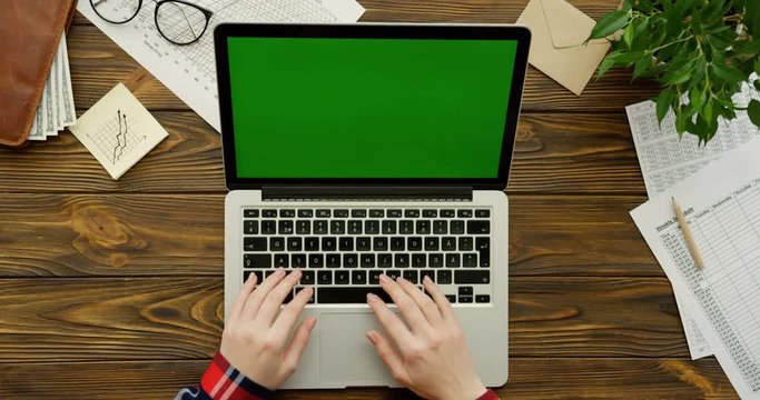 Close up of the female hands with the red plaid sleeves working on the laptop computer with a green screen on the wooden office table with office stuff and plant. Chroma key. Top view