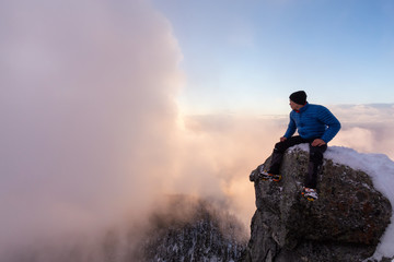 Adventurous man is sitting on the edge of a cliff and enjoying the beautiful mountain scenery. Taken in St Mark's Peak, North of Vancouver, British Columbia, Canada.
