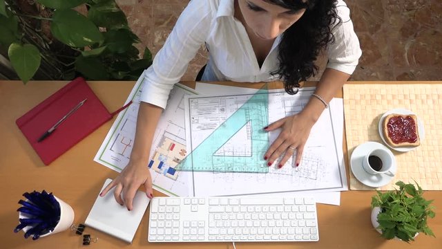 Female architect on desk drawing plan and having breakfast in the studio. Active engineer working hard at office proofreading floor plan, drinking coffee and eating bread and jam. Busy lifestyle