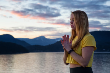 Fototapeta na wymiar Woman in prayer position is meditating with her eyes closed during a vibrant sunset. Taken in Whytecliff Park, Horseshoe Bay, West Vancouver, British Columbia, Canada.