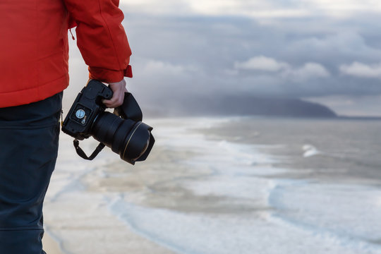 Man holding a camera in his hand overlooking the beautiful sandy beach during a vibrant winter sunrise. Taken in Cape Kiwanda, Pacific City, Oregon Coast, North America.