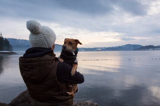 Young woman holding her little dog, Chihuahua, in her arms during a vibrant sunset. Taken in Belcarra, Vancouver, British Columbia, Canada.
