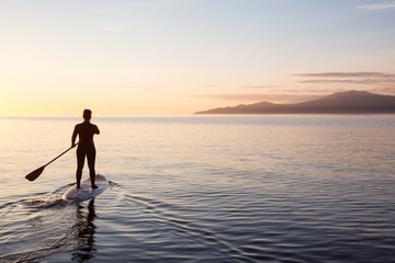 Adventurous girl on a paddle board is paddeling during a bright and vibrant sunset. Taken near...