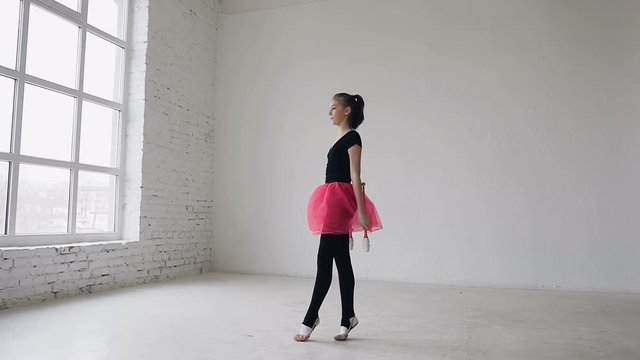 Cute gymnast doing circle with mace on the legs in the white studio on window background. Gymnastics girl dressed black bodysuit and pink skirt