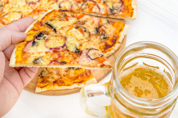 Hand holding piece of pizza over table with pizza and glass of beer.