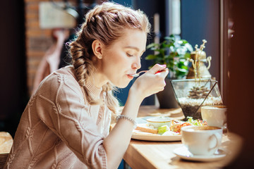 Charming woman with cute hairstyle having lunch during the rest in coffee shop, happy Caucasian female eating breakfast while relaxing in cafe during free time - obrazy, fototapety, plakaty