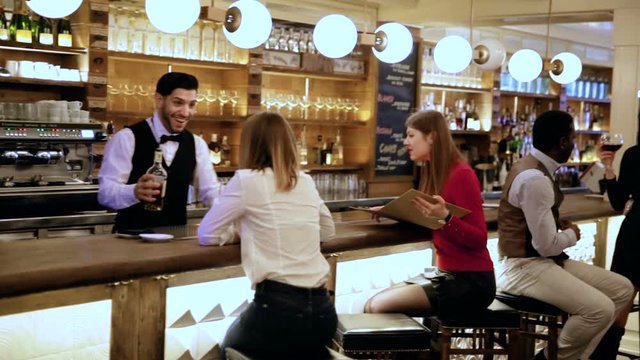 Smiling bartender pouring beverages and talking to cheerful visitors in bar
