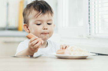 The child is sitting in the kitchen and greedily eats sausage