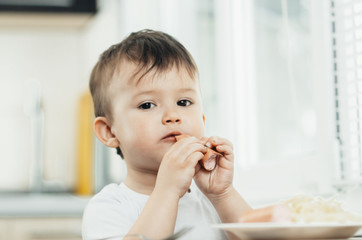 The child is sitting in the kitchen and greedily eats sausage