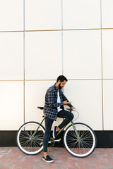Profile view of a trendy bearded hipster man using a mobile phone while sitting on bicycle, on the city street, near the urban wall. Dressed in shirt, jeans and moccasins.