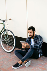 Fototapeta na wymiar Serious young bearded man using a computer tablet, reading or watching something, while sitting on the ground near the bicycle on the city street. Wearing plaid shirt and jeans.