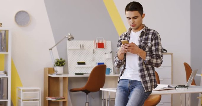 Young attractive brunette man scrolling and taping on the smartphone in the modern nice office while standing and leaning at the desk. Inside