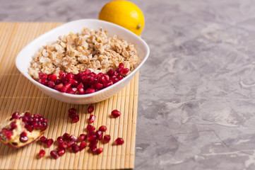 Cooked yummy warm oatmeal and heap of ripe fresh pomegranate seeds in white ceramic bowl near piece of pomegranate and whole lemon on clean bamboo rug on worn gray scratched concrete with copy space