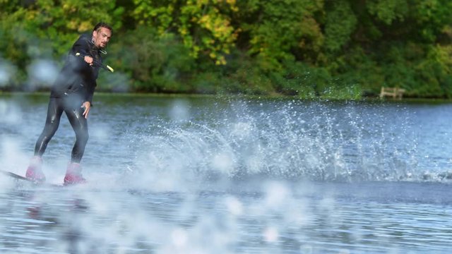 Handsome man wakesurfing in water splashes. Young man riding wakeboard on summer river. Waterskier moving fast in splashes. Wakeboarder surfing across river. Extreme lifestyle