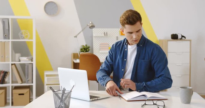 Young ood looking male startupper in the jeans shirt working on the laptop and writing in the notebook. Indoor