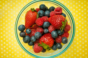 raspberries, strawberries and blueberries fruit in blue bowl over yellow background with copy space 