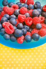 raspberries and blueberries fruit in blue bowl over yellow background with copy space 