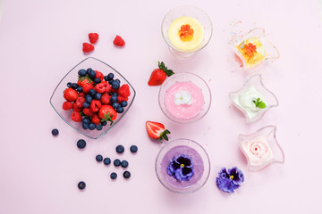 Ice cream in wave glass bowls and creamy milk beverage, smoothie with edible flowers *pansy, violet (on pink background. Pink (strawberry), yellow (mango or banana)  Top view