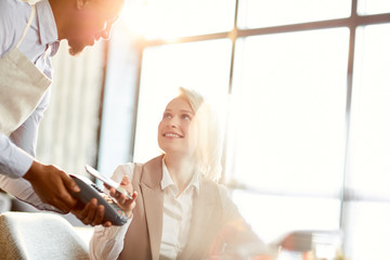 Happy contemporary woman looking at waiter while paying for order by her smartphone in cafe