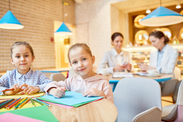 Pretty little girls with papers and crayons sitting by table in cafe while their mothers working by next table behind